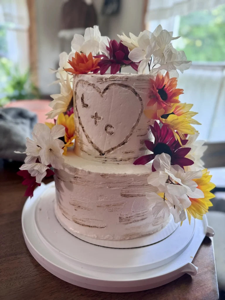 Two-tier white frosted cake with a heart illustration on the side, decorated with orange, yellow, and white flowers around the tiers.
